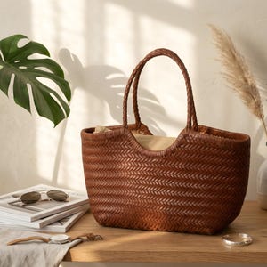 K&ouml;nnte beinhalten: Eine braune, gewebte Ledertasche mit geflochtenen Henkeln. Die Tasche hat eine rechteckige Form mit einer breiten &Ouml;ffnung. Der Innenraum ist mit einem hellfarbigen Stoff gef&uuml;ttert. Die Tasche steht auf einer Holzoberfl&auml;che.