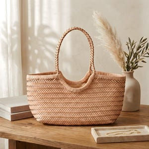 K&ouml;nnte beinhalten: Eine gewebte beigefarbene Tragetasche mit geflochtenem Griff. Die Tasche hat eine rechteckige Form und eine breite &Ouml;ffnung. Die Tasche steht auf einem Holztisch neben einer Vase mit dekorativen Pflanzen.