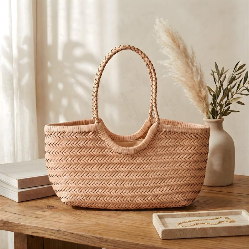 K&ouml;nnte beinhalten: Eine gewebte beigefarbene Tragetasche mit geflochtenem Griff. Die Tasche hat eine rechteckige Form und eine breite &Ouml;ffnung. Die Tasche steht auf einem Holztisch neben einer Vase mit dekorativen Pflanzen.