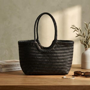 K&ouml;nnte beinhalten: Eine schwarze gewebte Tragetasche mit geflochtenen Griffen. Die Tasche hat eine rechteckige Form und eine strukturierte Oberfl&auml;che. Die Griffe sind gew&ouml;lbt und oben an der Tasche befestigt. Die Tasche steht auf einer Holzoberfl&auml;che.