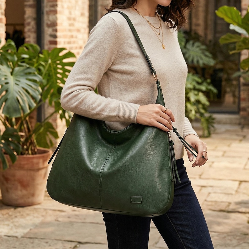 Puede incluir: Un bolso de hombro de cuero verde oscuro con un dise&ntilde;o curvo. El bolso tiene una correa larga y un detalle de borla. El bolso lo lleva una persona que lleva un su&eacute;ter beige y vaqueros azul oscuro.