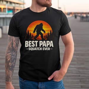Può includere: T-shirt nera con grafica di Bigfoot davanti a un tramonto. Il testo bianco "BEST PAPA - SQUATCH EVER" è stampato sotto. La maglietta è girocollo e viene indossata da una persona.