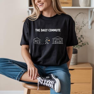 May include: Black t-shirt with the text "THE DAILY COMMUTE" above a line drawing of a house, person, and car in a garage. The person is walking with a coffee cup. The shirt is made of a soft material.
