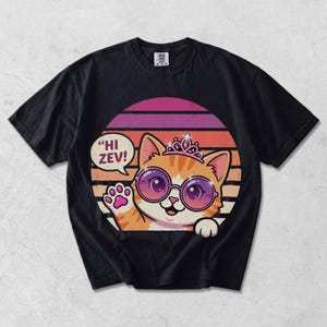 Könnte beinhalten: Schwarzes T-Shirt mit einer Cartoon-Katze, die eine Sonnenbrille und eine Tiara trägt und mit einer Pfote winkt. Die Katze ist orange und weiß, vor einem Retro-Sonnenuntergangsdesign mit dem Text "HI ZEV!" in einer Sprechblase.