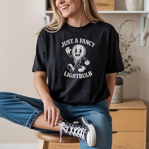 May include: Black t-shirt with the text "JUST A FANCY LIGHTBULB" and a cartoon lightbulb character. The shirt is worn with blue jeans and black and white sneakers. The person is sitting on a wooden stool.