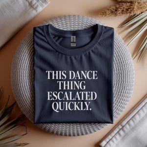 May include: Dark navy blue t-shirt featuring the white text "THIS DANCE THING ESCALATED QUICKLY". The shirt is presented on a circular, woven mat, with decorative elements in the background.
