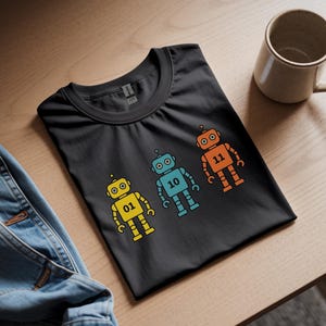 May include: Black t-shirt featuring three colorful robot illustrations. Each robot is a different color: yellow, blue, and orange, with the numbers 01, 10, and 11 on their chests. A beige mug sits nearby on a wooden surface.