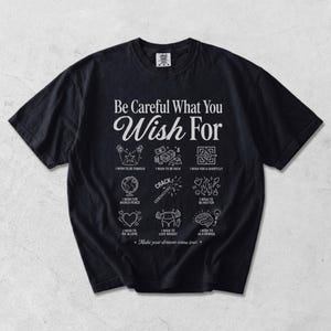 以下が含まれることがあります： Be Careful What You Wish Forという白いグラフィックテキストがプリントされた黒い半袖Tシャツ。テキストの下には、I Wish To Be Famous、I Wish To Be Rich、I Wish For A Shortcut、I Wish For World Peace、I Wish To Be Hotter、I Wish To Be In Love、I Wish To Lose Weight、I Wish To Be A Genius、Make your dreams come trueというラベルが付いた9つのアイコンが並んでいます。