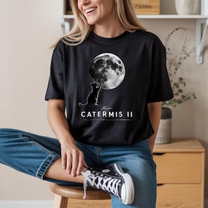 May include: Black t-shirt featuring a graphic of a cat reaching for a full moon. The text "Meow... CATERMIS II" is printed below the cat and moon design. The shirt is a crew neck style.