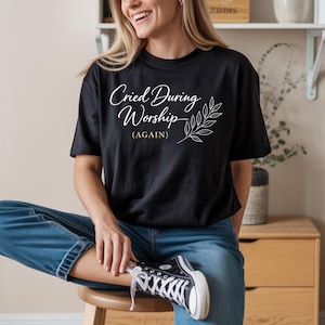 Puede incluir: Camiseta negra con el texto "Cried During Worship (AGAIN)" en fuente cursiva, acompañada de un diseño de hojas. La persona lleva vaqueros azules y zapatillas altas negras.