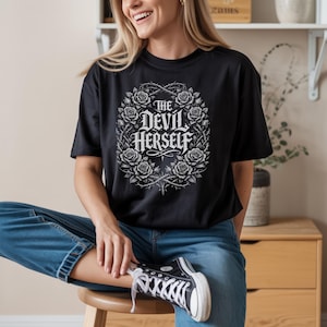 Puede incluir: Camiseta negra con el texto "The Devil Herself" en fuente gótica, rodeada por una corona de rosas y espinas. La camiseta se combina con vaqueros azules y zapatillas deportivas blancas y negras.