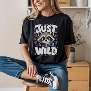 Puede incluir: Una camiseta negra de manga corta con un gráfico de una cara de mapache y el texto "JUST A LITTLE WILD" en letras blancas y azules, rodeado de pequeñas estrellas, corazones y hojas decorativas en colores rojo, blanco y azul.