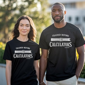Puede incluir: Dos personas con camisetas negras con el texto blanco "TRAINED BEFORE CALCULATORS". Las camisetas presentan un gráfico de una regla de cálculo y el texto "EST. PRE 2010". El fondo está borroso, con vegetación y luz solar.