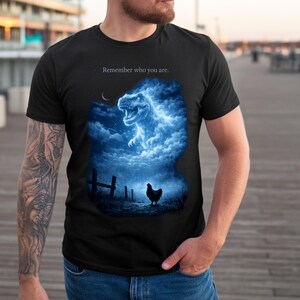 Puede incluir: Camiseta negra con un gráfico azul y blanco de un T-Rex en una formación de nubes, una gallina y el texto "Remember who you are." El diseño está ambientado en un cielo nocturno.