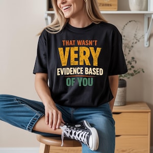 Könnte beinhalten: Schwarzes T-Shirt mit dem Text "THAT WASN'T VERY EVIDENCE BASED OF YOU" in Orange, Gelb und Grün. Das Shirt wird mit blauen Jeans und schwarz-weißen Turnschuhen getragen. Die Person sitzt auf einem Holzschemel.