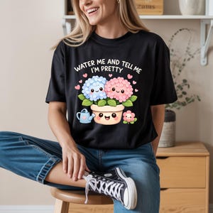 Puede incluir: Camiseta negra con el texto "WATER ME AND TELL ME I'M PRETTY" sobre una ilustración de dibujos animados de dos flores en macetas con caras sonrientes. También se muestra una pequeña regadera y una flor más pequeña.