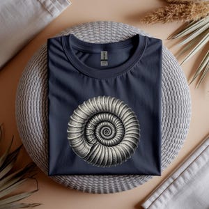 Puede incluir: Camiseta azul marino con una ilustración detallada en blanco y negro de una concha de nautilus. El gráfico está centrado en la camiseta. La camiseta se muestra sobre una superficie redonda y tejida con elementos decorativos.