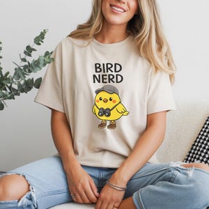 May include: Beige t-shirt with the words "BIRD NERD" in black block letters. Below the text is a cartoon yellow bird wearing a gray cap, binoculars, and brown boots. The shirt is worn by a person.