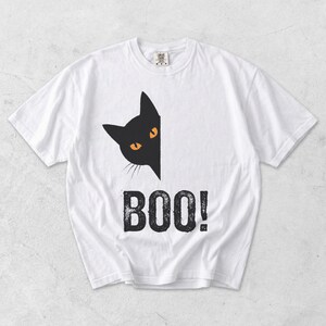 May include: White t-shirt featuring a black cat graphic with orange eyes peeking out from the side. Below the cat is the word "BOO!" in a distressed, bold font, perfect for Halloween.