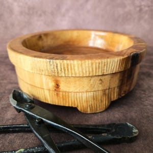 Puede incluir: Un cuenco de madera con un borde ancho y un exterior tallado se encuentra junto a un par de tijeras de bonsái negras. El cuenco está hecho de madera clara con un interior más oscuro. Las tijeras son de metal.
