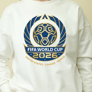 May include: White sweatshirt with the FIFA World Cup 2026 logo. The design features a blue and gold soccer ball graphic, the text "FIFA WORLD CUP 2026", and the host nations: United States, Canada, and Mexico.
