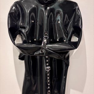 Könnte beinhalten: Eine schwarze, glänzende Latex-Zwangsjacke hängt an einem Metallbügel. Das Kleidungsstück hat einen hohen Kragen, mehrere silberfarbene Druckknöpfe und eine Reihe von Schnallen und Riemen. Die Ärmel sind über der Brust befestigt.