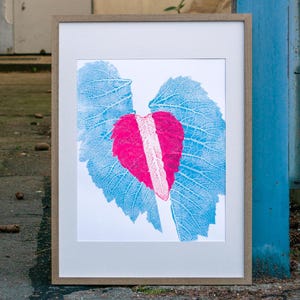 May include: Framed artwork featuring a heart-shaped design with a pink center and blue leaf-like wings. The artwork is in a light brown frame and leans against a blue structure. The background includes a door and a fence.