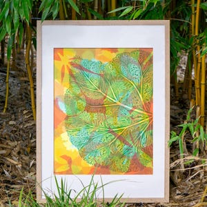 Puede incluir: Una lámina enmarcada que muestra una hoja verde grande y detallada con venas prominentes, superpuesta sobre un fondo vibrante de formas abstractas amarillas, naranjas y rojas. La obra está en un marco de madera clara con un paspartú blanco.
