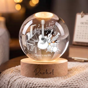 May include: A clear glass sphere lamp featuring an etched design of a guitar, piano, saxophone, drums, and musical notes with the text "Music is my life". The sphere rests on a cylindrical wooden base engraved with the name "Rachel".