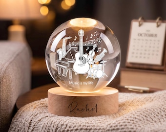 May include: A clear glass sphere lamp featuring an etched design of a guitar, piano, saxophone, drums, and musical notes with the text "Music is my life". The sphere rests on a cylindrical wooden base engraved with the name "Rachel".