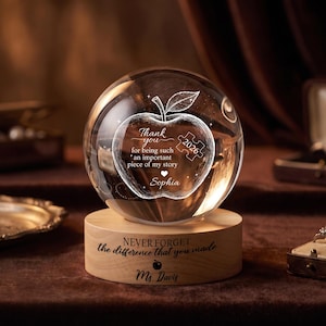 May include: A clear glass sphere engraved with an apple and a puzzle piece containing the text 'Thank you for being such an important piece of my story' and 'Sophia'. It sits on a cylindrical wooden base inscribed with 'NEVER FORGET the difference that you made' and 'Ms Davis'.