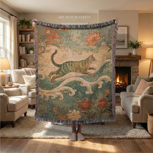Può includere: Una coperta tessuta con un disegno di un gatto che salta sopra onde e fiori nei toni del verde, beige e ruggine. La coperta ha un bordo con frange e il testo "ART MUSE BLANKETS" in alto.