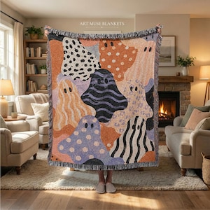 Peut inclure: Un plaid tissé présentant un motif répétitif de silhouettes de fantômes dans diverses couleurs et textures, incluant des pois, des rayures et des lignes ondulées. La couverture possède des bords à franges et le texte ART MUSE BLANKETS en haut.