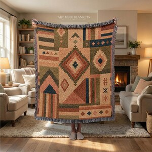 May include: A woven throw blanket featuring a geometric patchwork design in shades of terracotta, sage green, and navy blue. The blanket displays various patterns including diamonds, triangles, and stripes with a fringed edge and the text ART MUSE BLANKETS.
