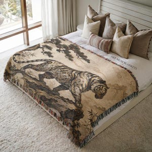 May include: A woven throw blanket featuring a large tiger illustration in shades of brown, black, and cream. The design includes stylized clouds, pine trees, and bamboo stalks. The text ART MUSE BLANKETS is printed in the corner of the fabric.