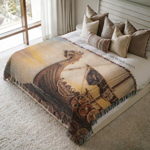 May include: A woven throw blanket featuring a detailed depiction of a Viking longship with a dragon-headed prow, circular shields along the side, and a sail. The design includes a golden sunset background and the text ART MUSE BLANKETS.