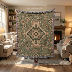 Peut inclure: Un plaid tissé présentant un fond vert sauge avec un grand motif central en losange et des motifs géométriques plus petits en rouille, crème et bleu foncé. La couverture a un bord à franges et le texte ART MUSE BLANKETS en haut.