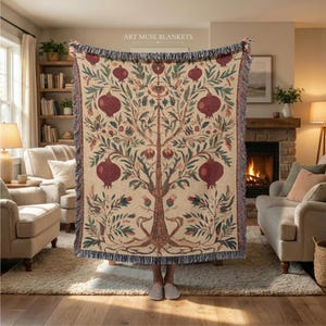May include: A woven throw blanket featuring a repeating botanical pattern of dark red pomegranates, green leaves, and brown branches on a light beige background. The blanket has a fringed edge and the text ART MUSE BLANKETS at the top center.