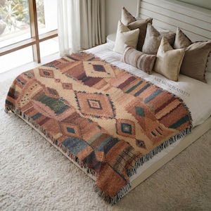 May include: A woven throw blanket featuring a geometric pattern in shades of rust, tan, blue, and green with fringe edges. The blanket displays the text ART MUSE BLANKETS. It is draped over a bed with several neutral-colored pillows and white bedding.