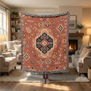 May include: A woven throw blanket in rust orange, cream, and dark blue featuring a central geometric medallion and scattered tribal-inspired motifs. The blanket has a fringed edge and the text ART MUSE BLANKETS printed at the top centre.
