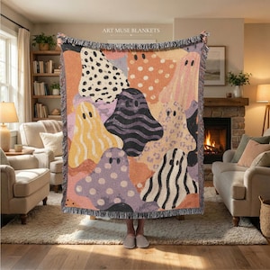May include: A woven throw blanket featuring a repeating pattern of ghost-shaped figures in various colours and textures, including polka dots, stripes, and leopard print, against a warm-toned background with fringed edges and the text ART MUSE BLANKETS.