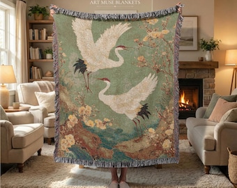 May include: A woven throw blanket featuring a sage green background with two white cranes in flight, red crown markings, and yellow floral accents. The blanket has a fringed edge and the text ART MUSE BLANKETS printed at the top center.