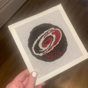 May include: A square piece of white needlepoint canvas featuring a circular black hockey puck design with a red and white swirling storm logo in the center. The number 2026 is stitched in red thread at the bottom of the circular puck graphic.