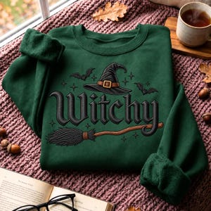 May include: A dark green long-sleeve sweatshirt featuring a graphic print of a black witch hat, a brown broomstick, and two small bats. The text "Witchy" is printed in a bold, dark font across the center, surrounded by small star accents.