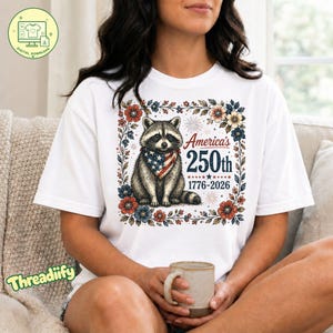 Peut inclure: T-shirt blanc avec un raton laveur portant un bandana patriotique, entouré de fleurs. Le design comprend le texte "America's 250th 1776-2026". Le t-shirt est un téléchargement numérique.