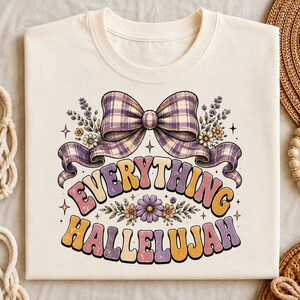 Peut inclure: Un t-shirt couleur crème avec une impression graphique représentant un grand nœud à carreaux dans des tons violets et beiges, entouré de petites fleurs et de brins de lavande. Le texte "EVERYTHING HALLELUJAH" est imprimé dans une police rétro.