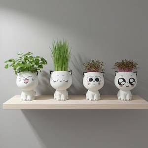 Op de afbeelding: Vier witte keramische katvormige plantenbakken met verschillende gezichtsuitdrukkingen, elk met een andere plant. Ze staan op een lichtgekleurde houten plank tegen een grijze achtergrond. De planten omvatten groene bladplanten en hoog groen gras.