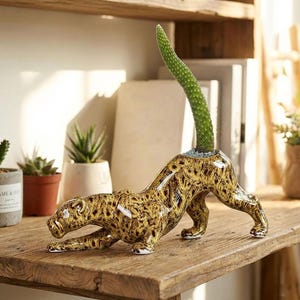 May include: A ceramic leopard-shaped planter with a green cactus growing from its back. The leopard features a brown and yellow spotted pattern. The planter is displayed on a wooden shelf.