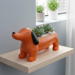 May include: Orange dachshund-shaped planter with black ears and a smiling face. The planter holds two green succulents in a bed of small white stones. The planter is on a light wood shelf.