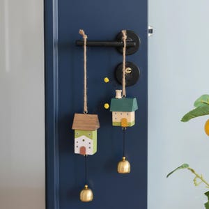 May include: Two miniature wooden house ornaments hanging from twine, each with a small gold bell. The houses are painted in green, white, and brown, with small windows and doors. They are hanging on a dark blue door.